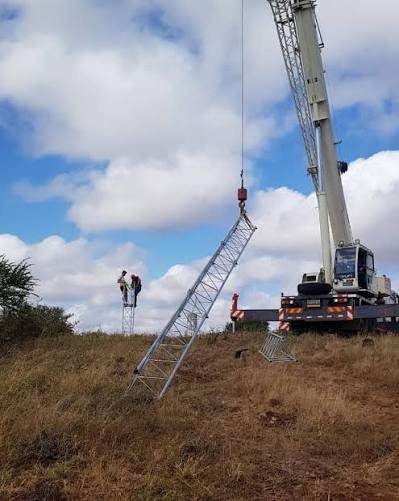 Crane lifting a galvanised lattice tower section toward climbers on a hilltop mast