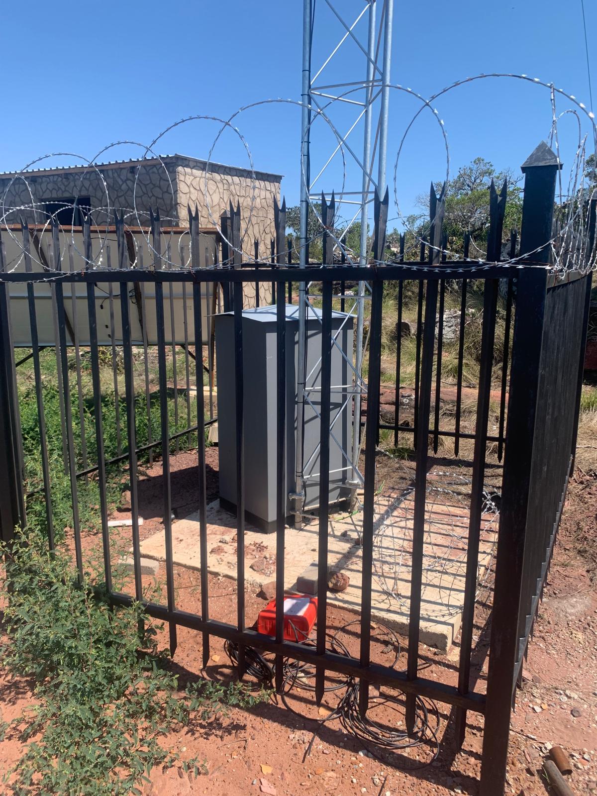 Grey outdoor equipment cabinet inside a palisade fence with razor wire at a tower site